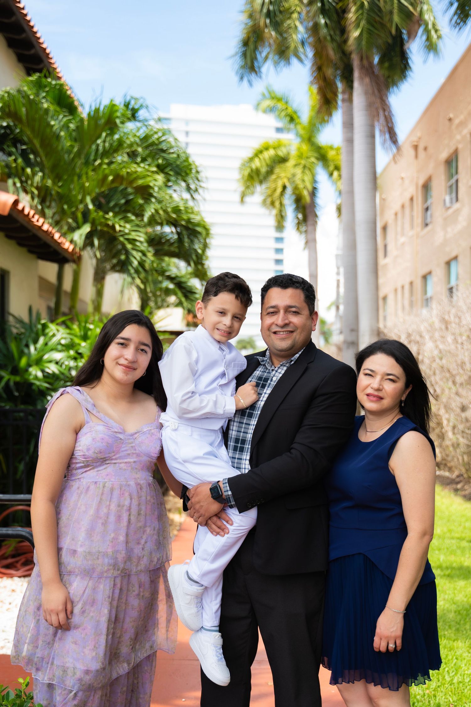 Family of four posing outdoors with palm trees, wearing formal attire including suits and dresses, smiling together.