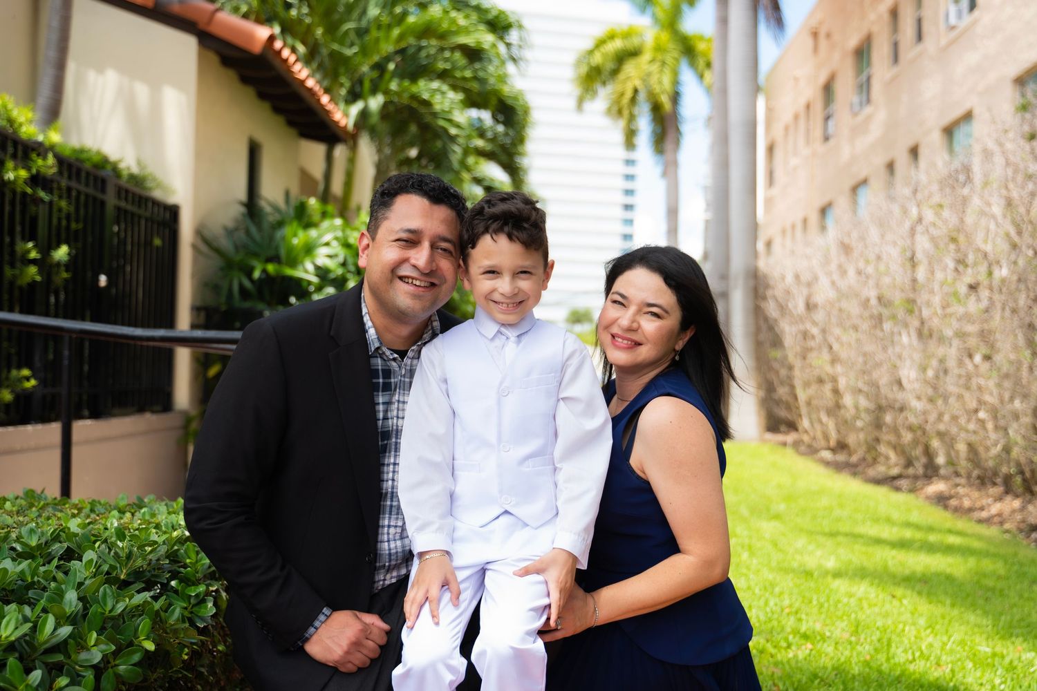 A happy family of three poses outdoors, parents holding young boy in white outfit, lush greenery background.