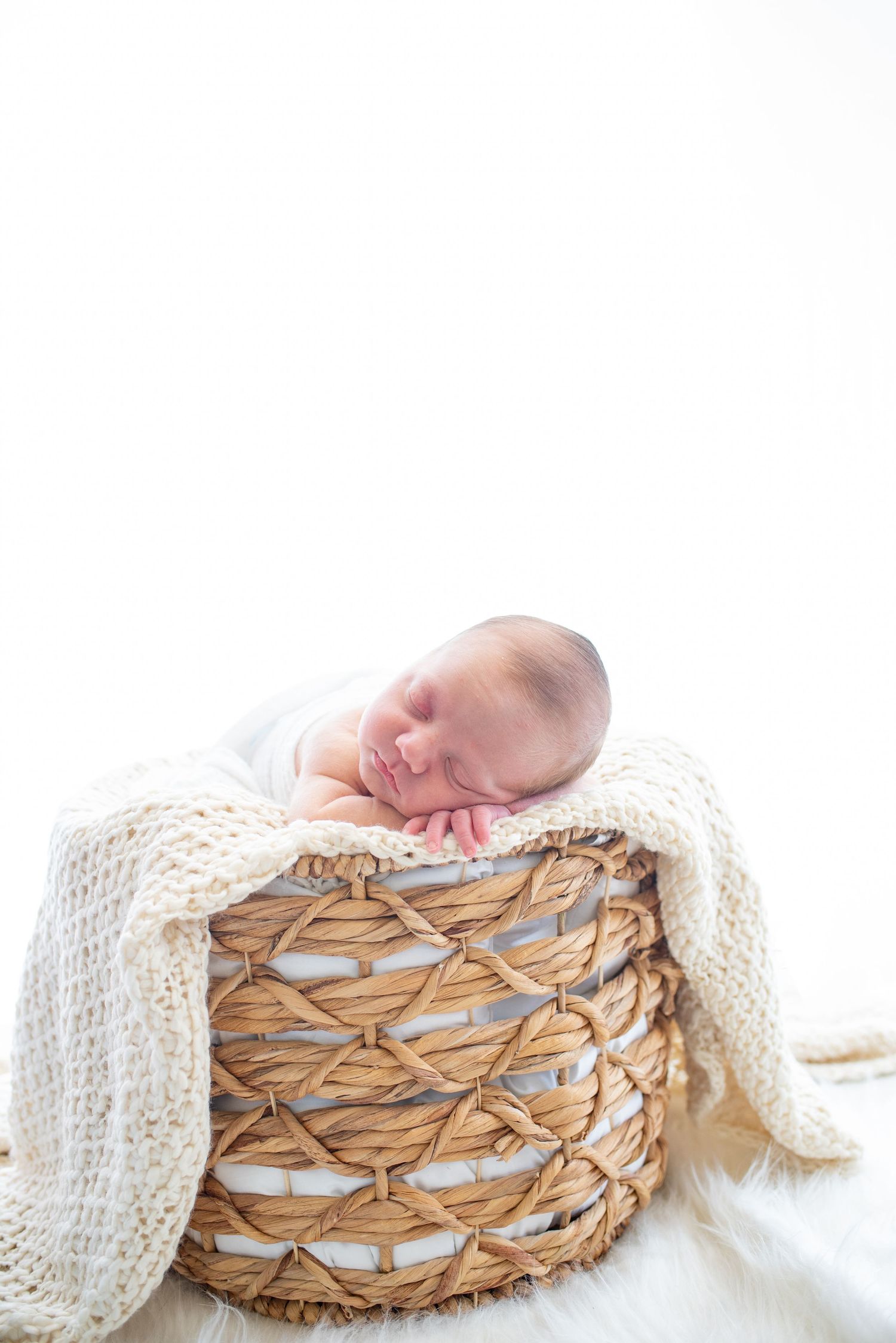Newborn sleeping peacefully in a woven basket draped with white knit blanket.