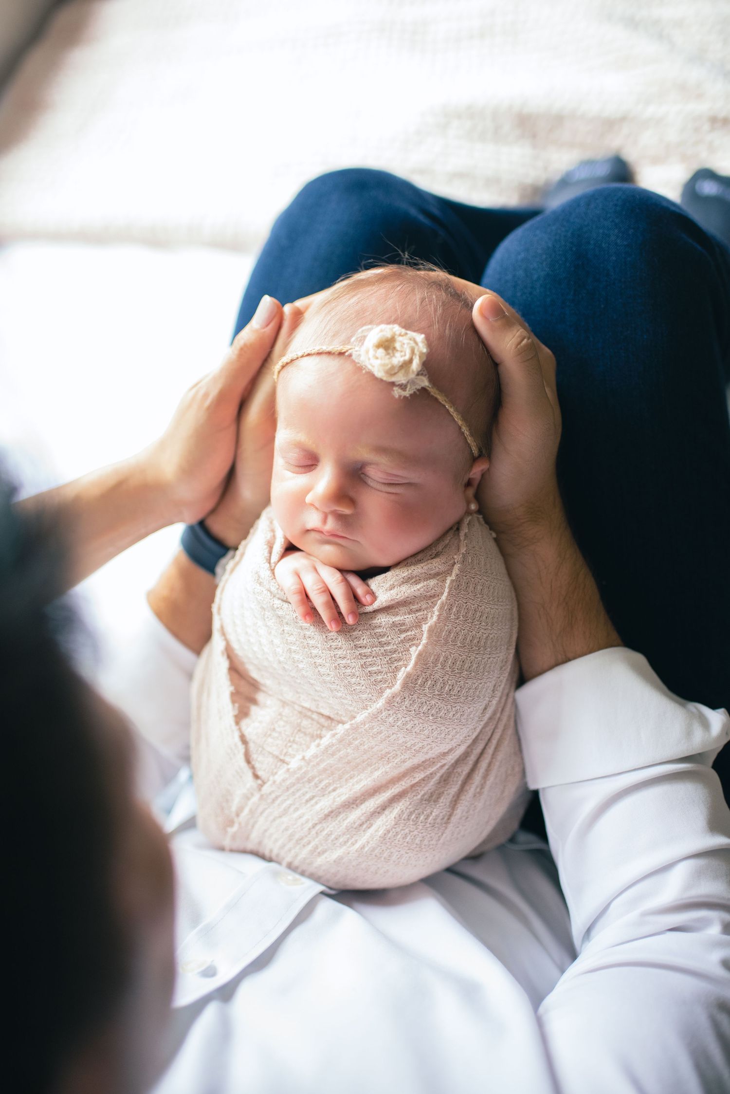 A sleeping newborn wrapped in soft beige fabric with a delicate headband being cradled by gentle hands.