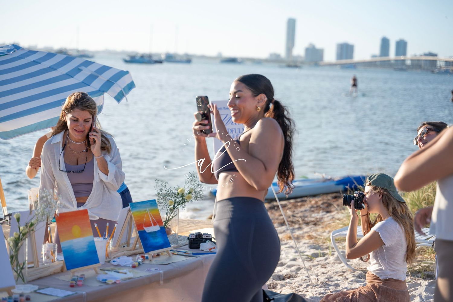 Group enjoying a picnic spread with drinks and snacks on a sunny beach with city skyline in background.