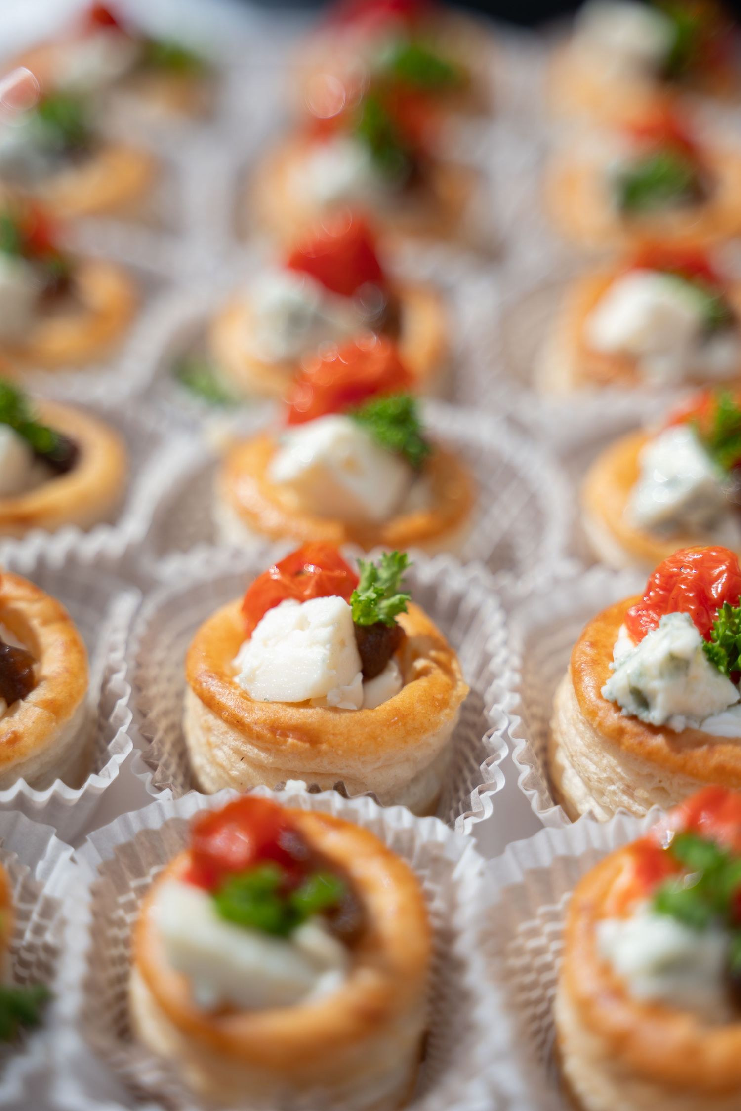Small appetizer bites with cream filling and red tomato garnish arranged in rows on white paper liners.