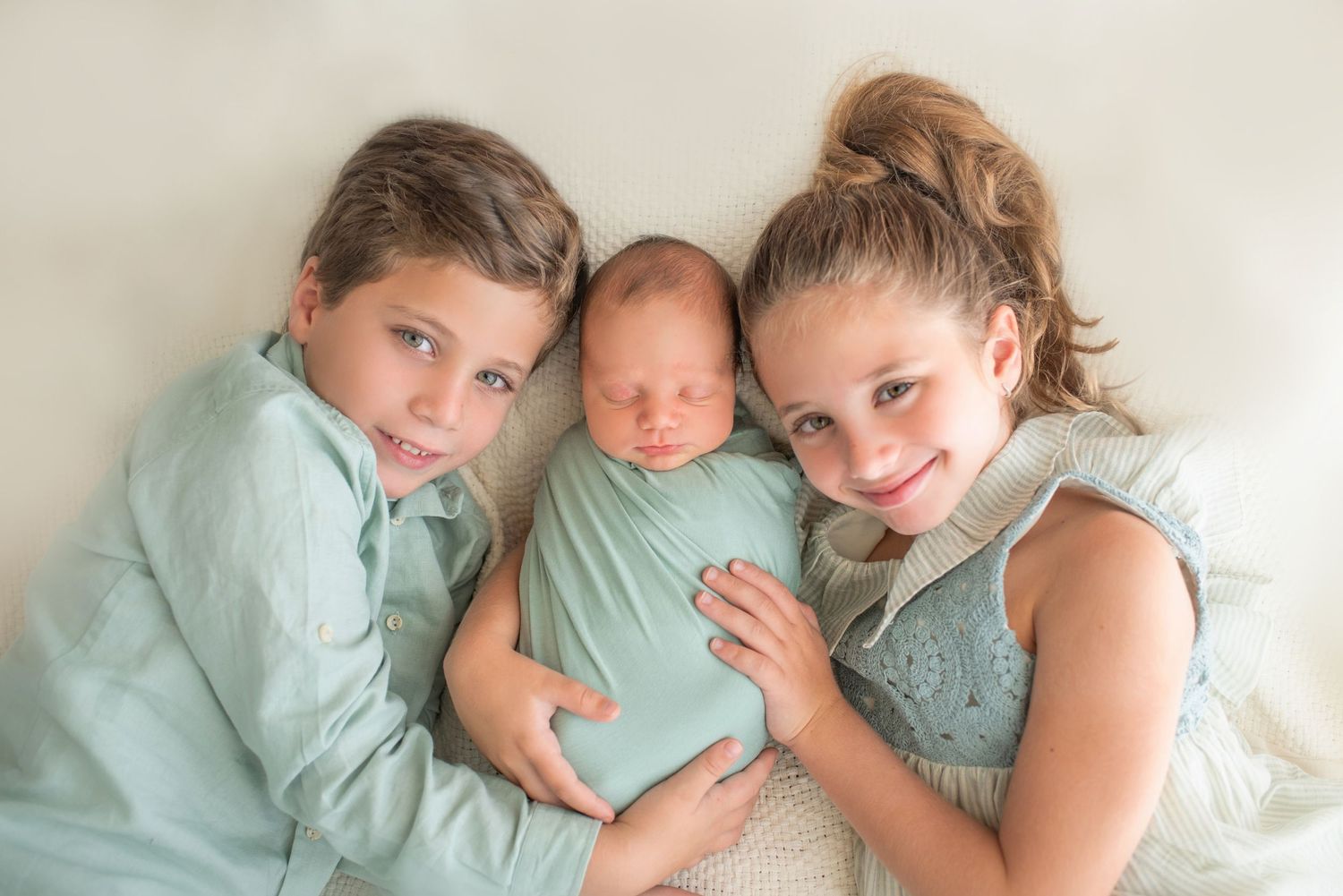 Three siblings in soft mint green outfits cuddle together on a white background in a tender family portrait.