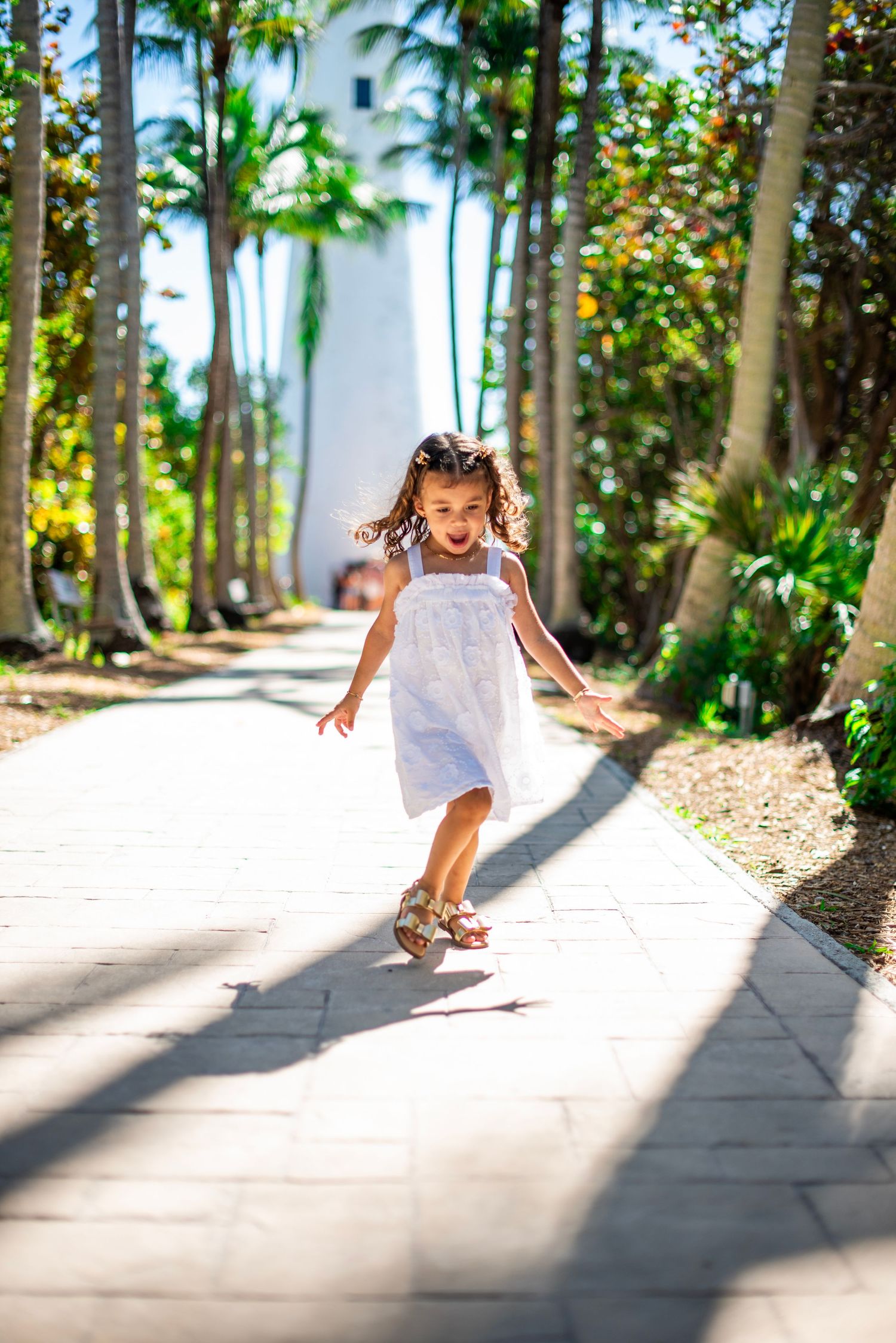 A child in a white dress skips playfully along a palm tree-lined pathway in bright tropical sunlight.