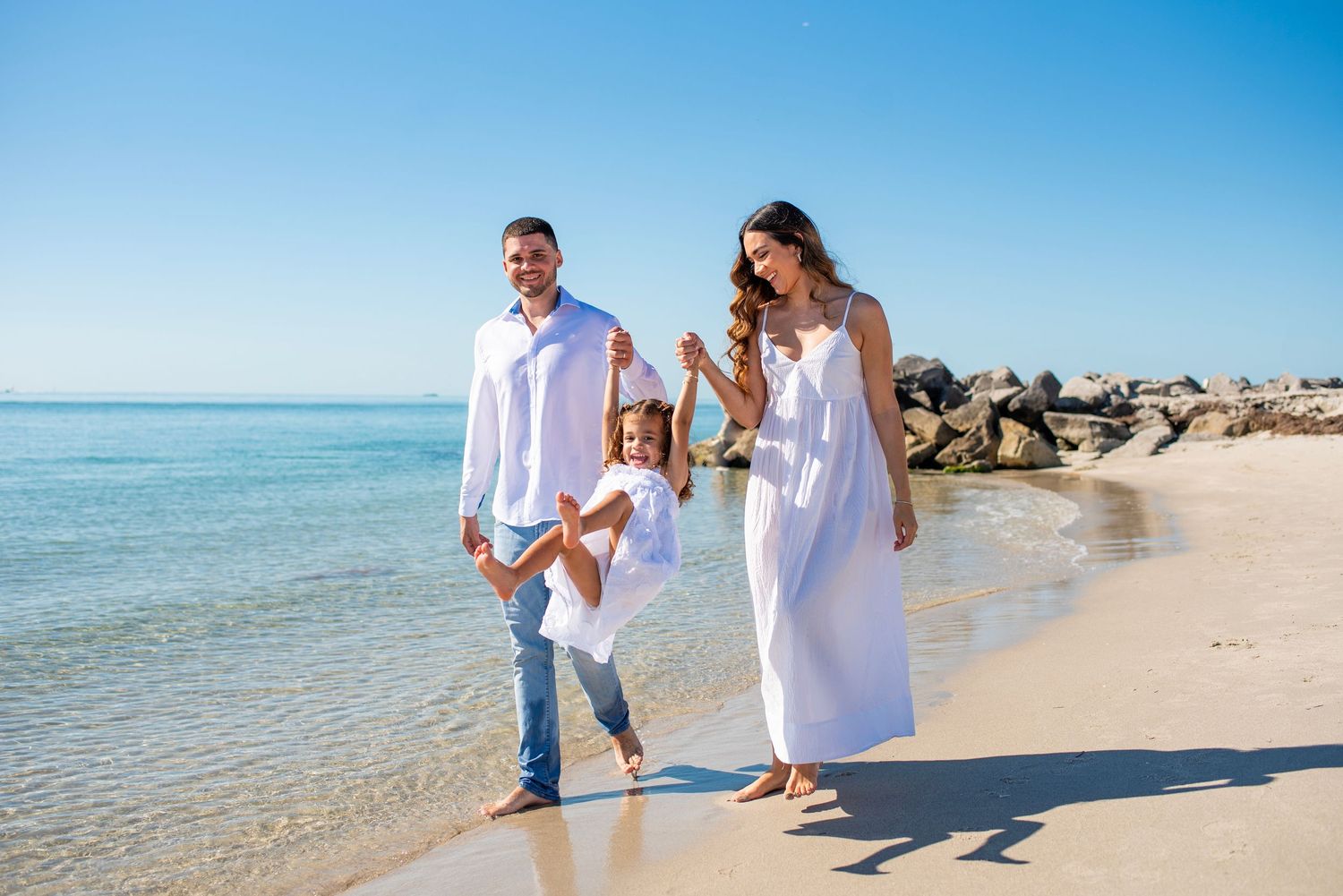 A happy family walks together along a pristine white sand beach wearing white outfits against a turquoise ocean backdrop.