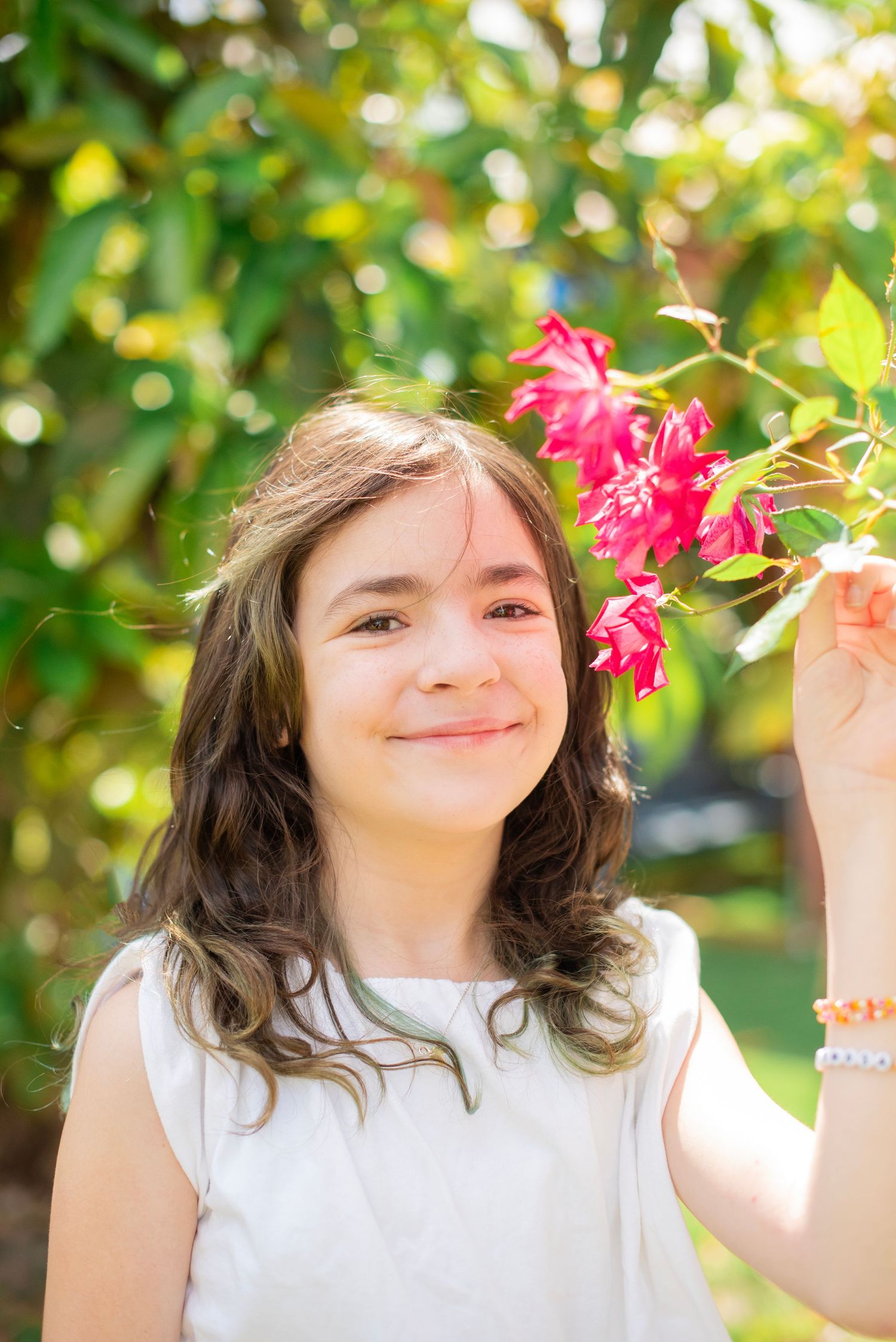 A young person in a white sleeveless top poses outdoors near pink flowers with natural bokeh in the background.