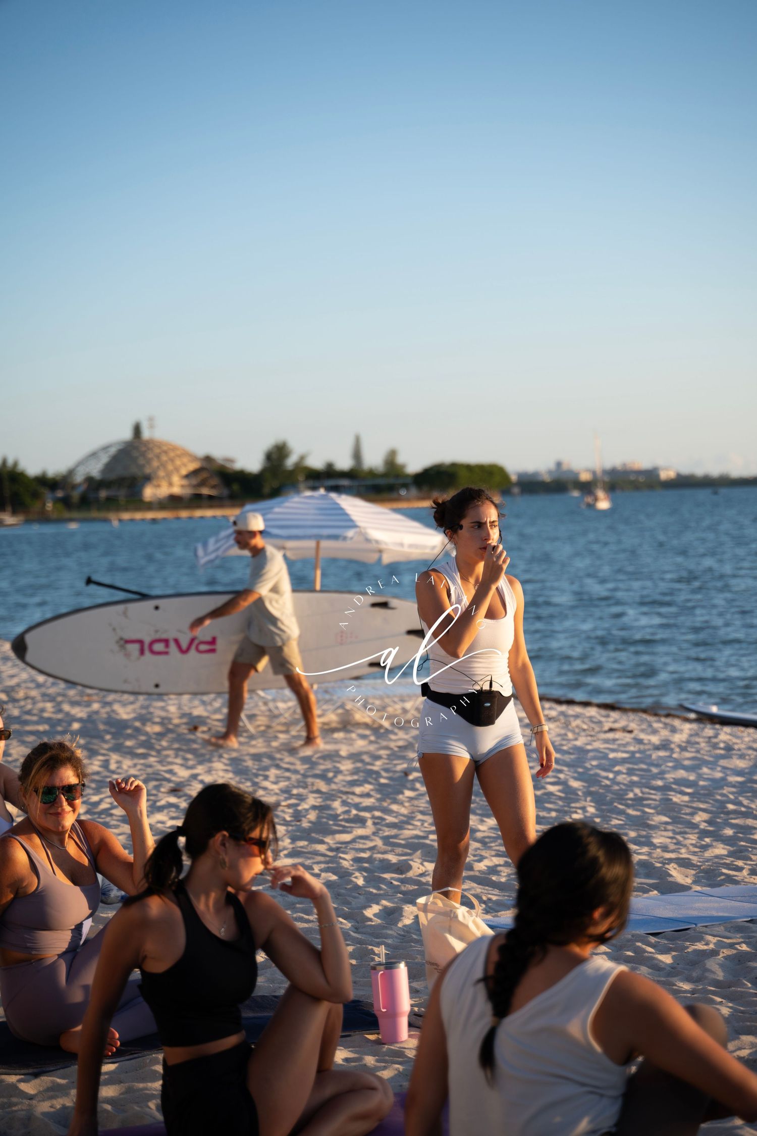 A group in swimwear relaxes on a sandy beach at sunset with paddleboards and drinks nearby.
