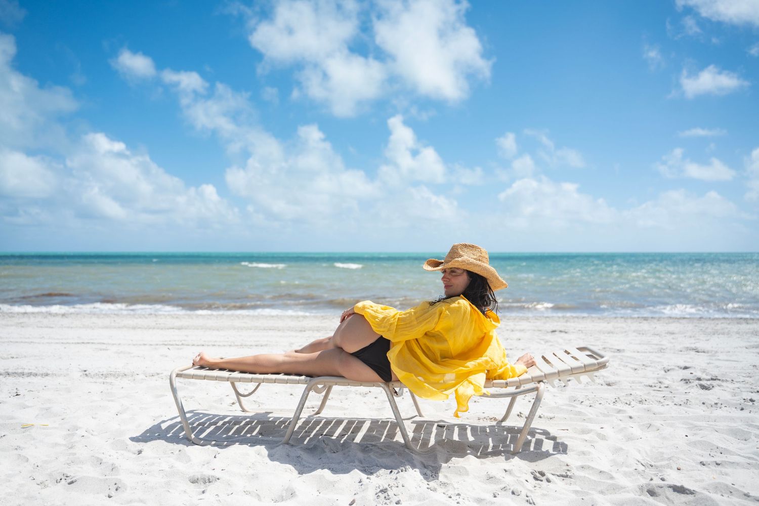 Someone in a yellow dress and sun hat relaxes on a beach lounger by the turquoise ocean on a sunny day.
