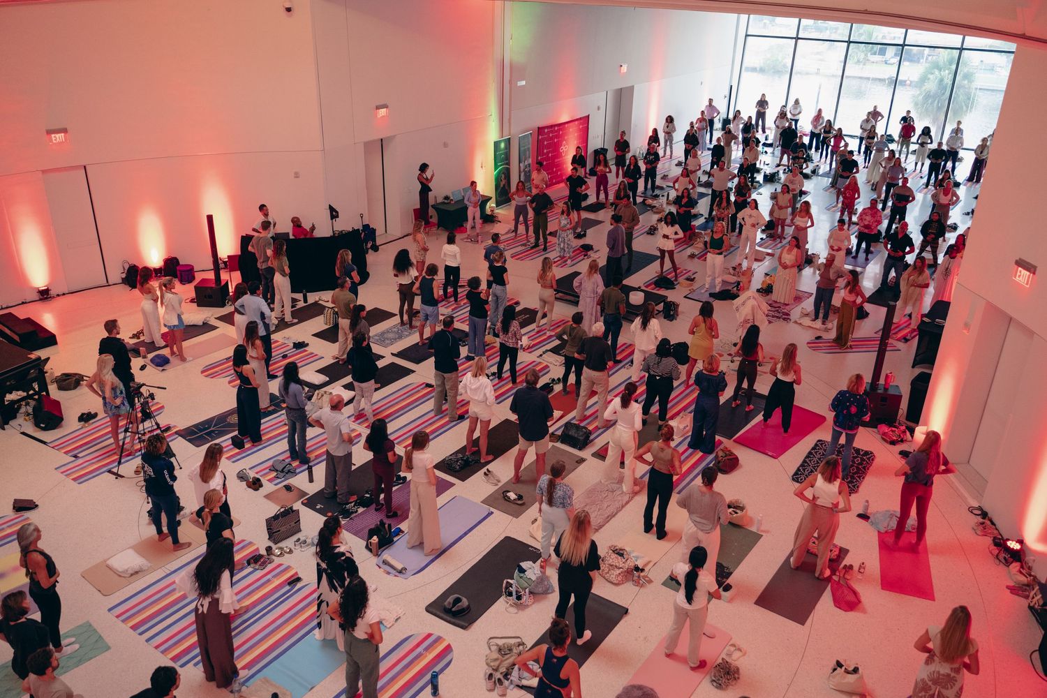 Large group yoga class in a bright modern studio with colorful mats and ambient pink lighting.