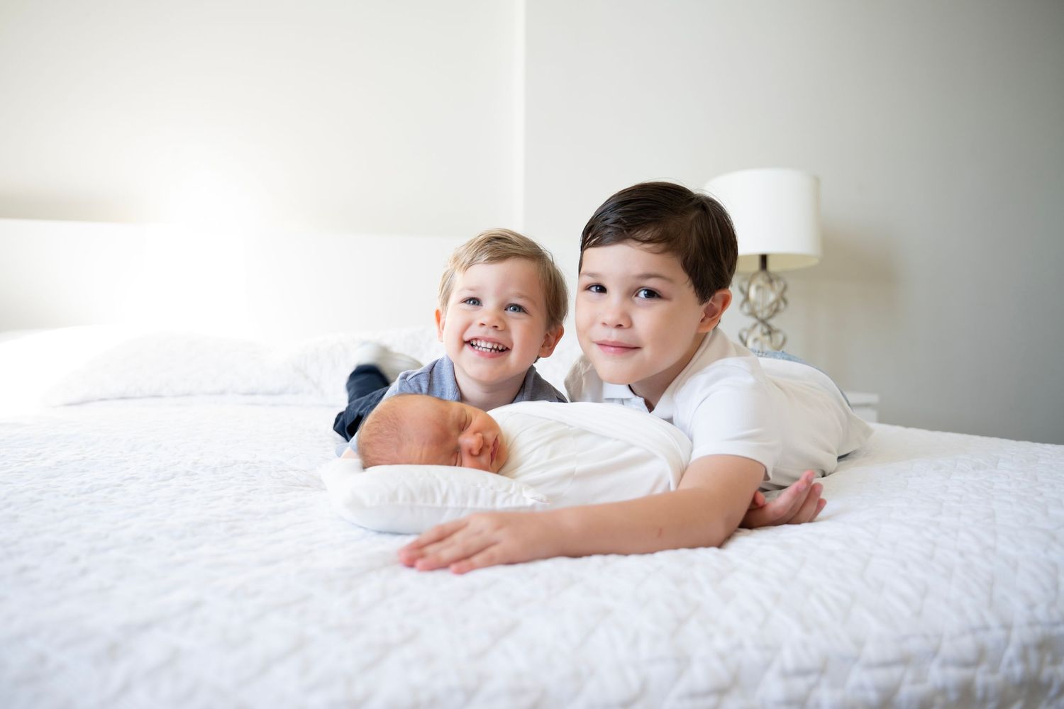 Two children lying on a white bed with pillows, smiling and relaxing together.