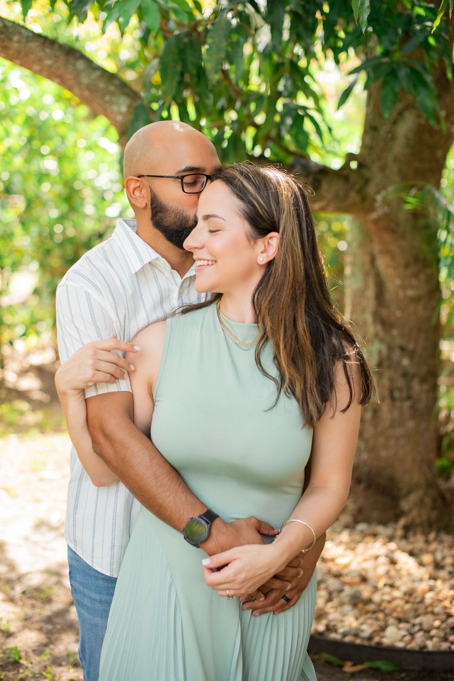 A couple embraces lovingly under green trees in an outdoor setting during a romantic photoshoot.
