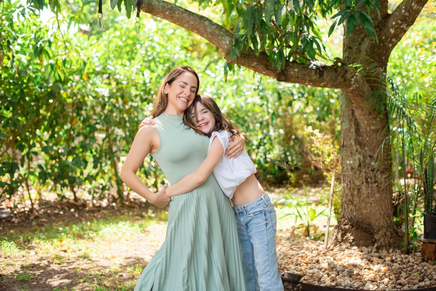 A couple in light blue and white outfits embraces under a tree in a sunny orchard setting.