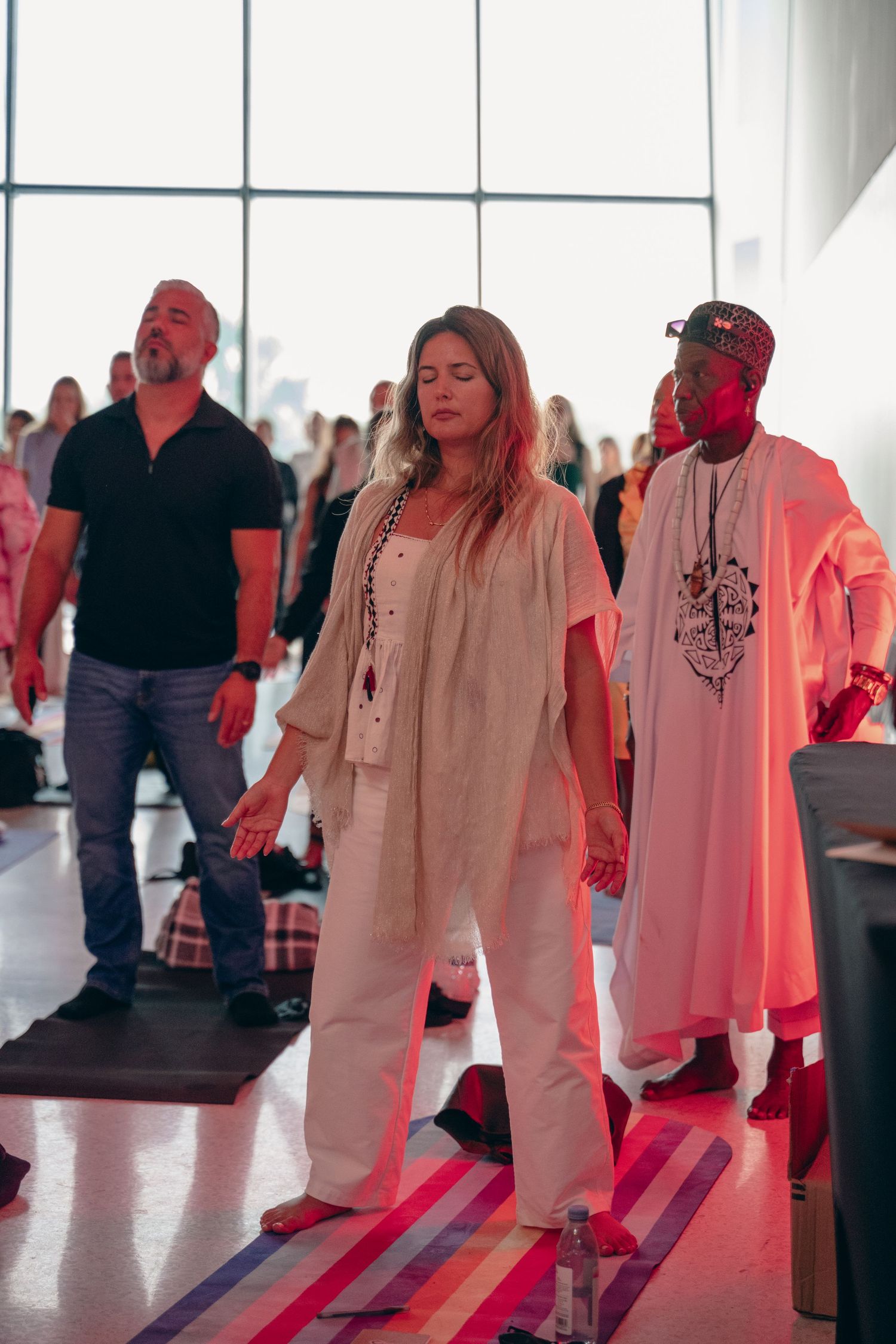 People in white attire stand meditating with eyes closed in a bright, modern event space with colorful floor lighting.