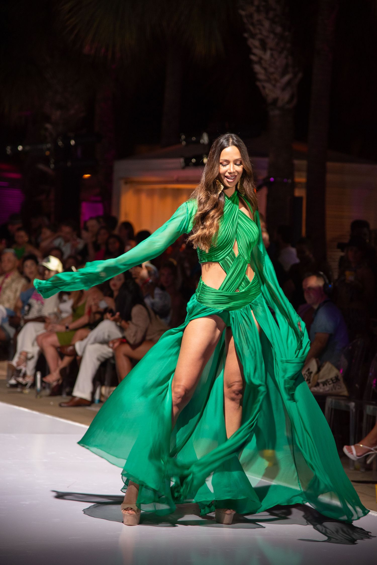 Model twirls in flowing emerald green cutout gown on fashion runway with palm trees in background.