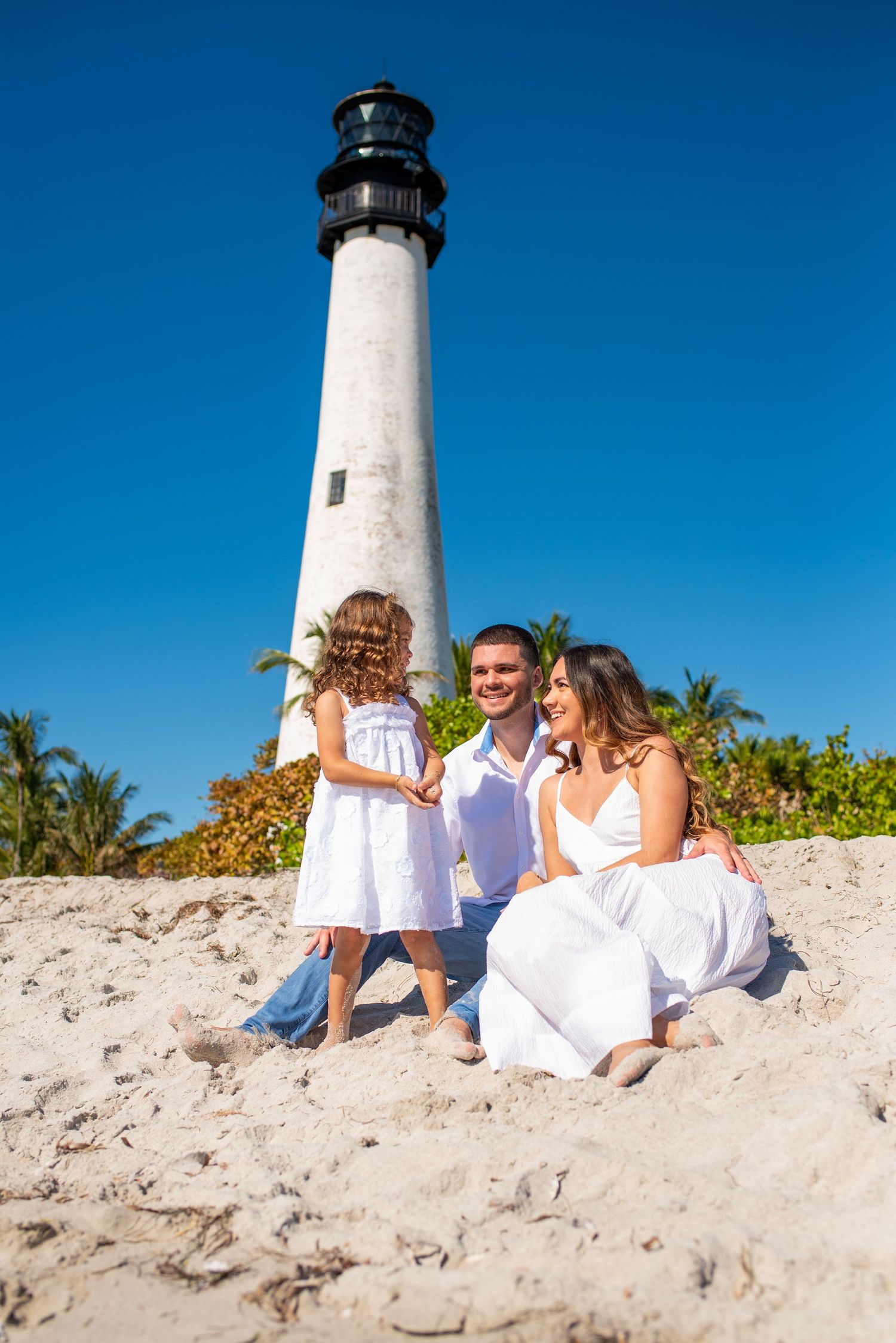 A white lighthouse tower stands tall against bright blue sky with palm trees and sandy beach in Key Biscayne, Florida.