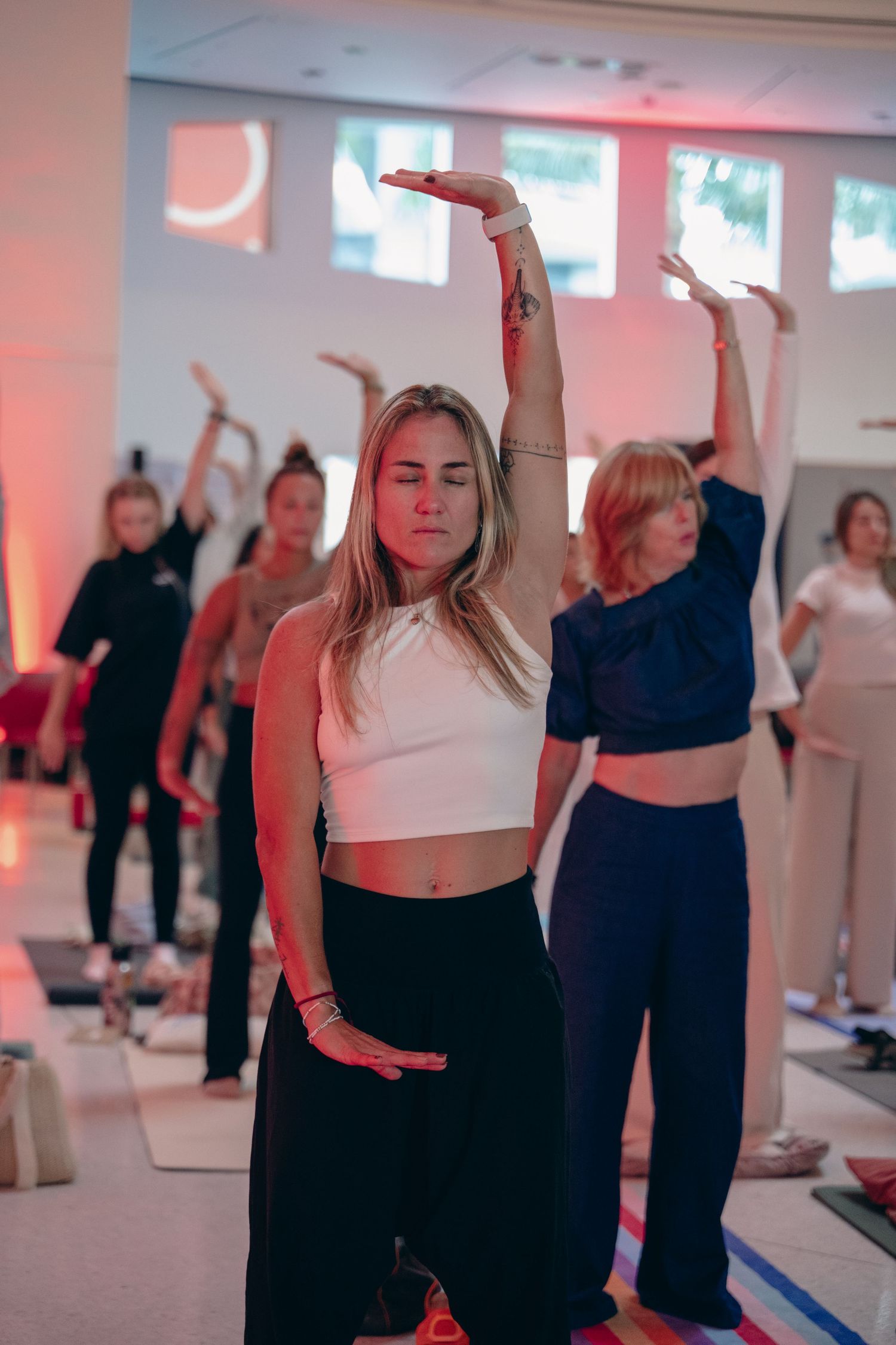 Women practicing yoga in a studio with warm red lighting, raising arms in synchronized movement during a group class.