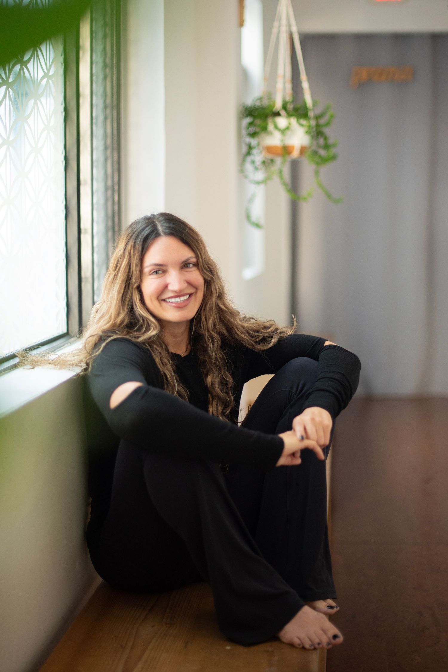 Person in black sweater poses playfully near window with hanging plants and natural lighting in modern space.