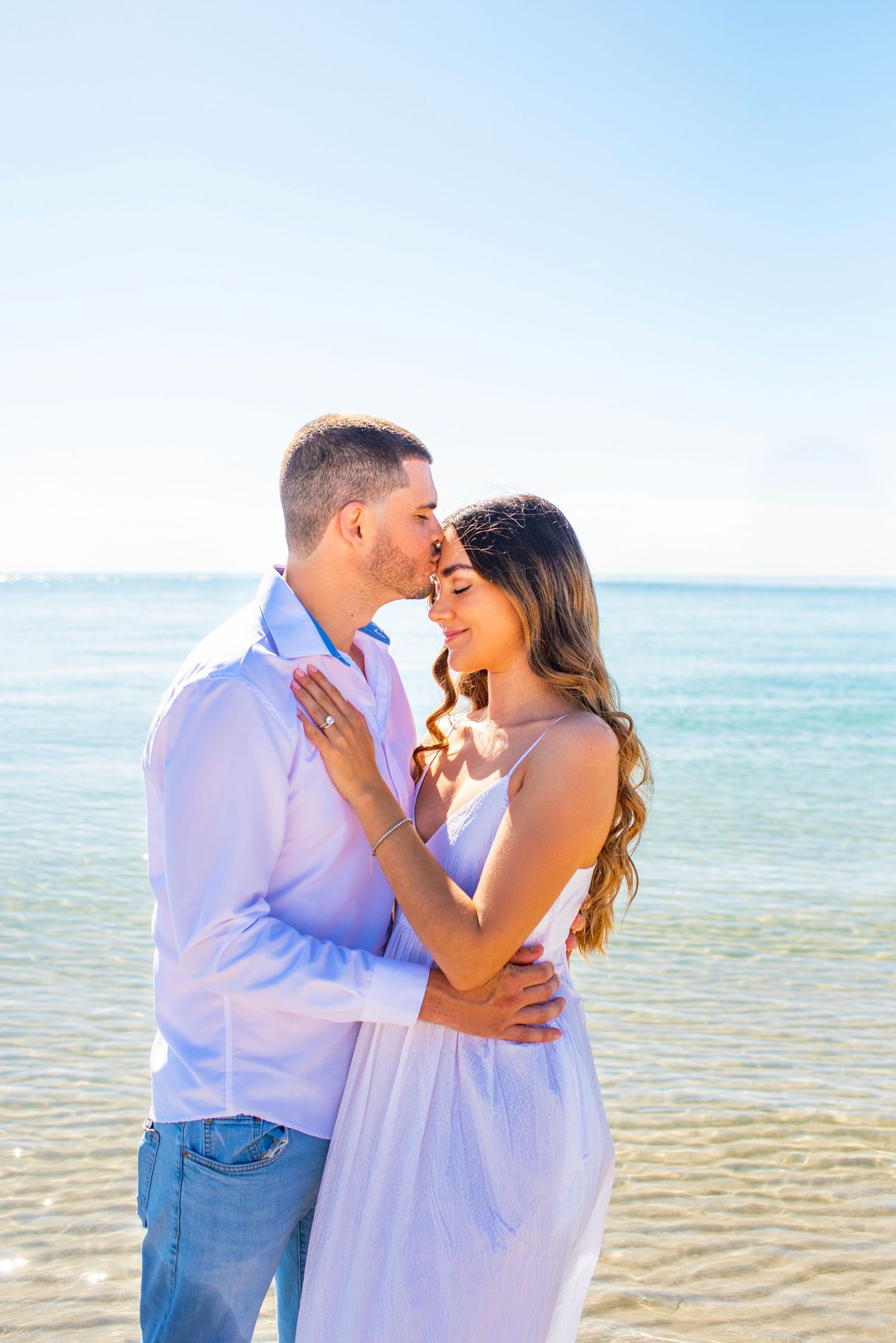 A romantic couple embraces on a sunny beach with crystal blue water and clear skies in the background.