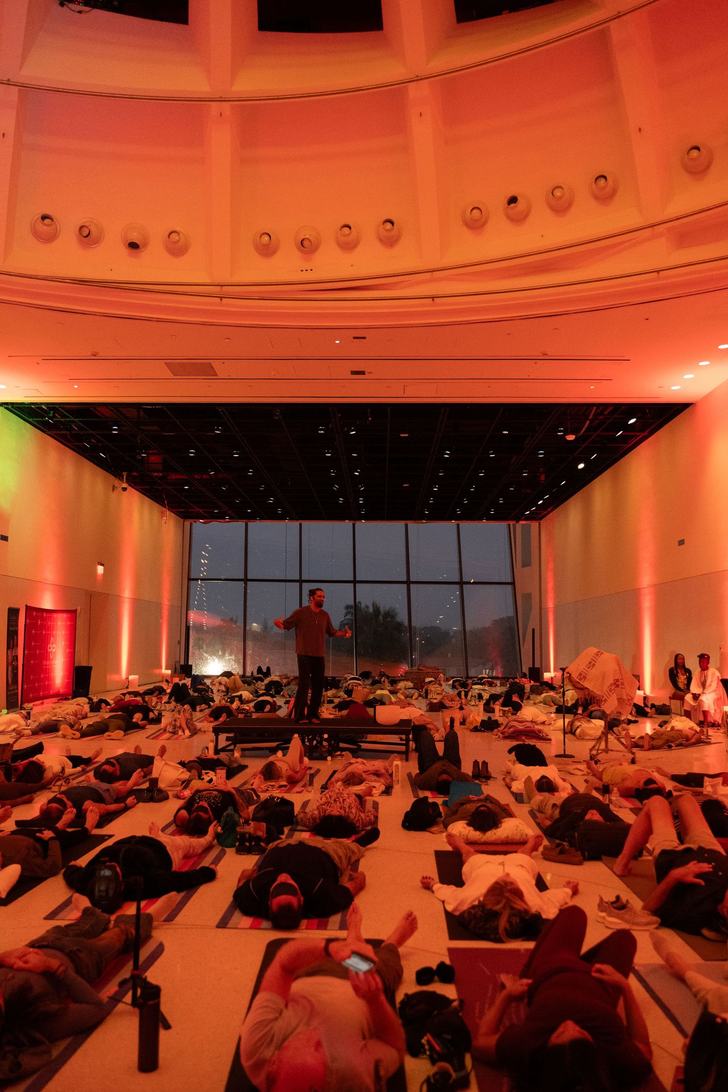 Large group of people lying on floor in grand hall with orange ambient lighting and floor-to-ceiling windows.