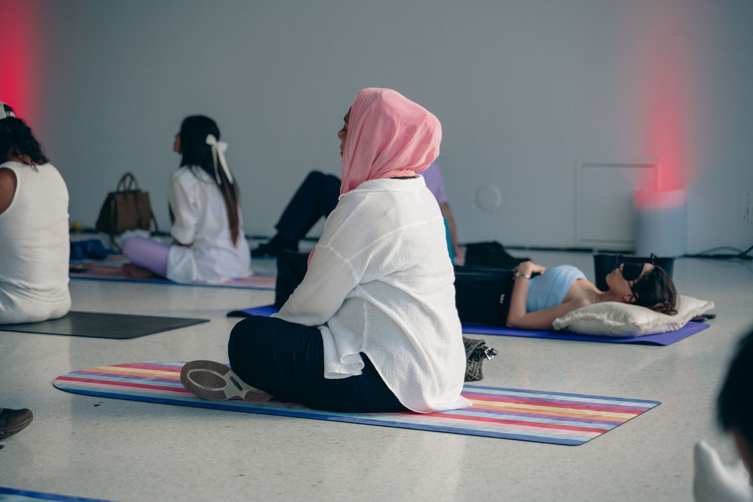Women practicing yoga on colorful mats in a bright studio, one wearing a pink hijab seated in a resting pose.
