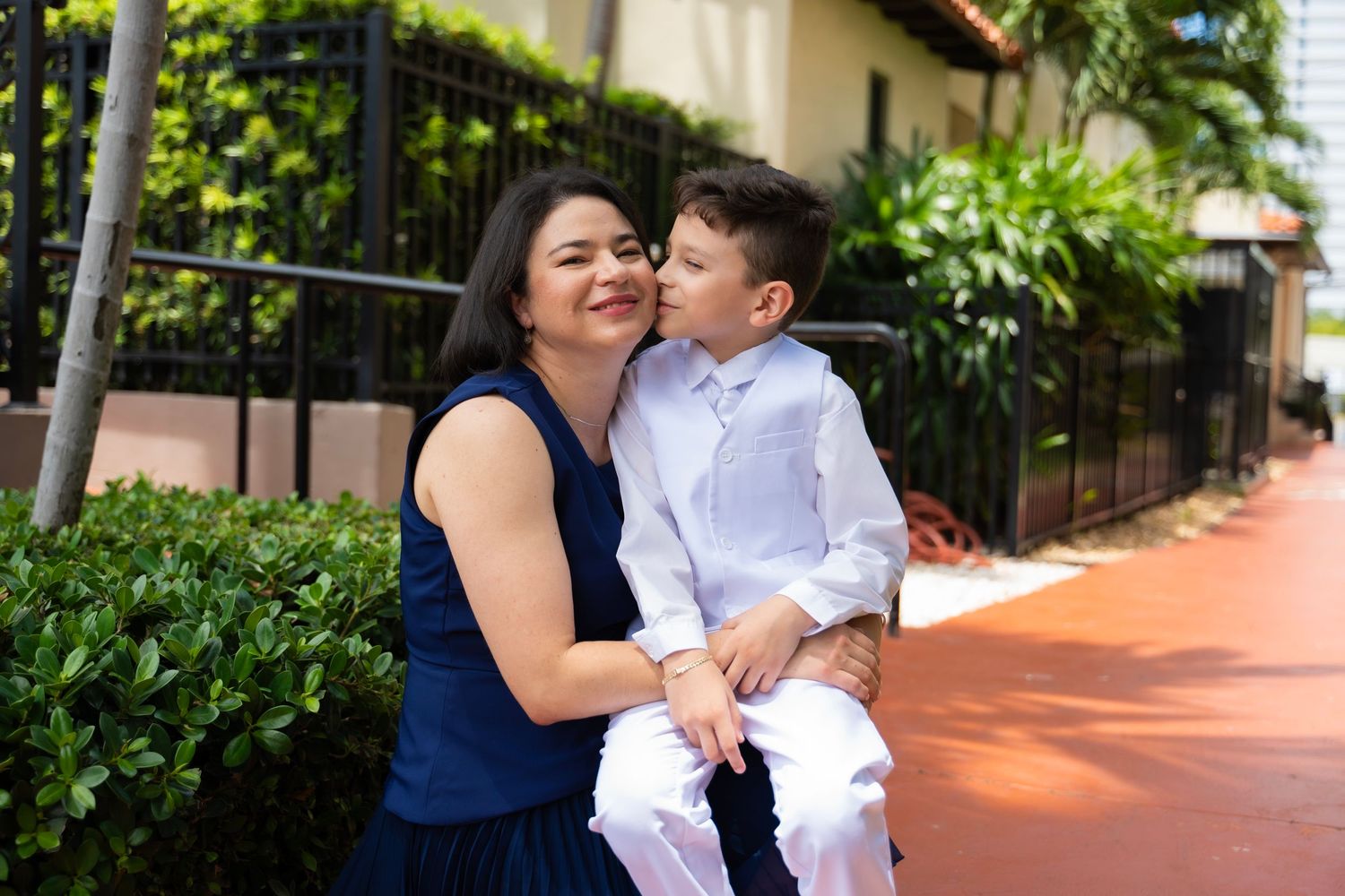 A woman in a blue dress holds a young boy in white who kisses her cheek outdoors.