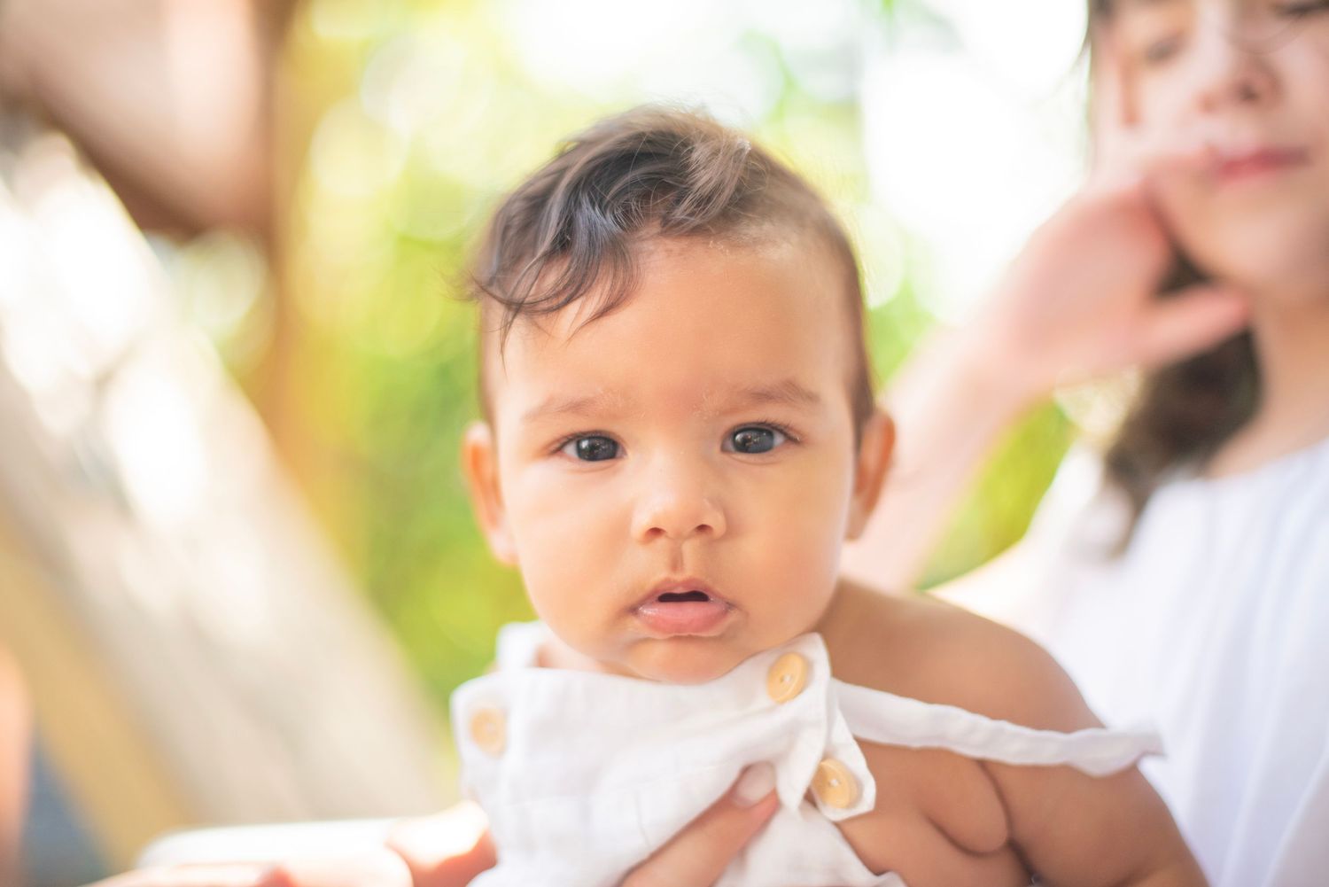 A baby in a white dress smiles while being held outdoors in natural lighting.