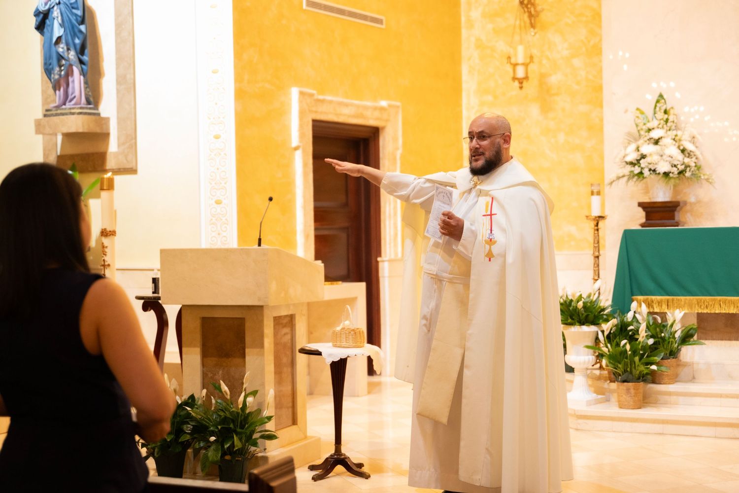 A priest in white vestments gestures during a Catholic church ceremony at an altar with yellow walls and floral arrangements.