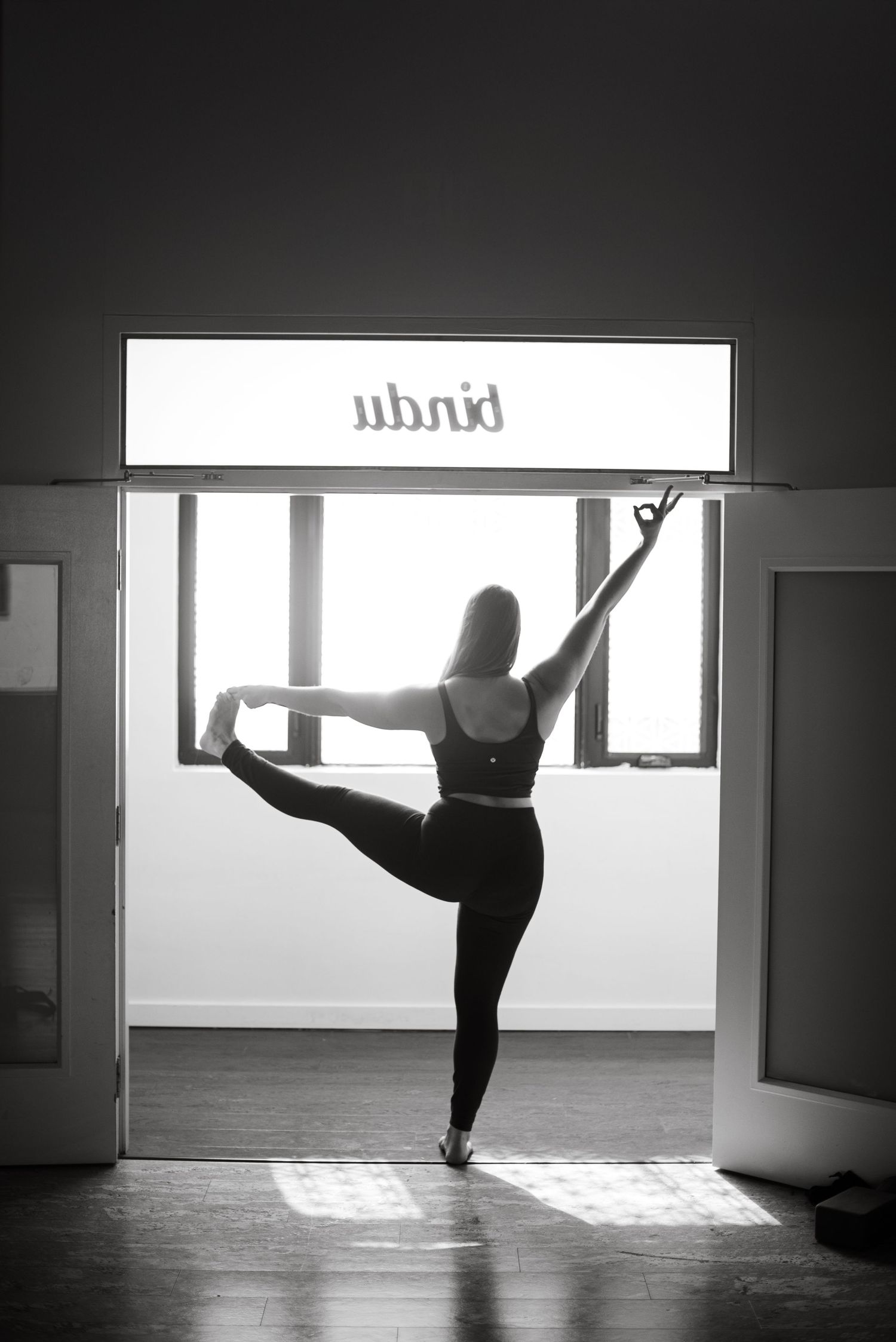 A dancer performs a graceful arabesque pose in silhouette against a bright window in a dance studio.