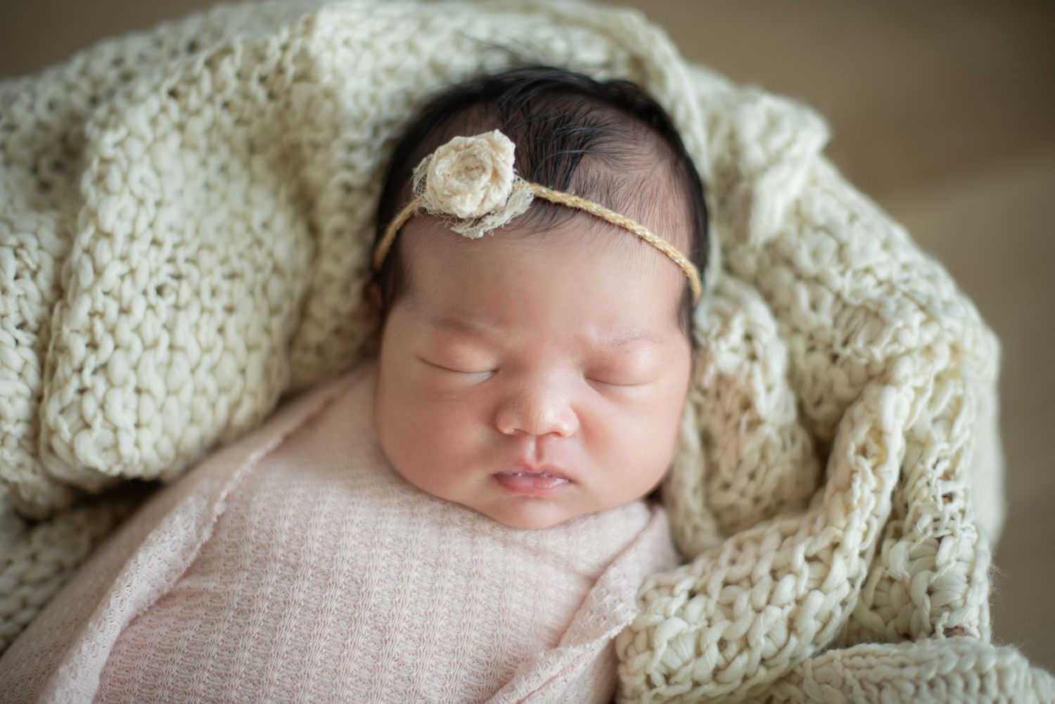 A sleeping newborn baby wrapped in cream-colored knit blankets with a delicate lace headband rests peacefully.