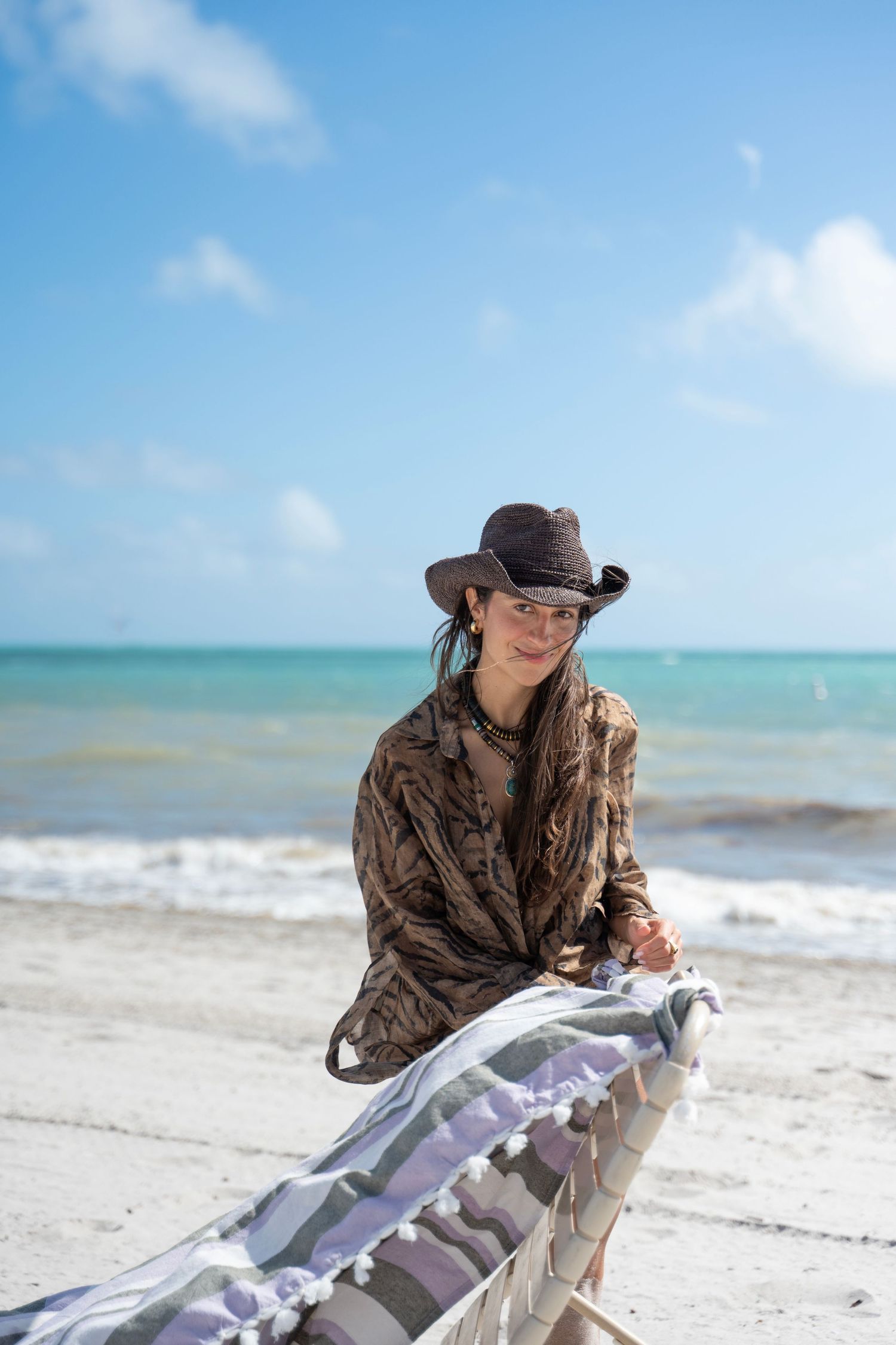 Person in brown clothing and hat lies on white sandy beach with turquoise ocean waters and blue sky in background.