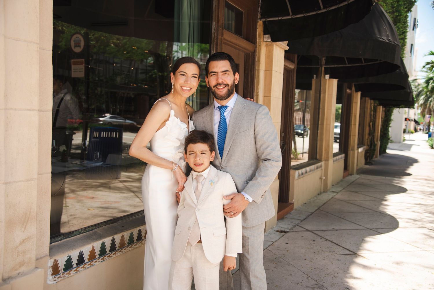 Wedding party members share embraces along a covered walkway with palm trees.
