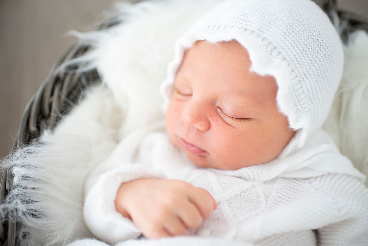 Newborn baby wearing white knit hat sleeps peacefully on white textured blanket.