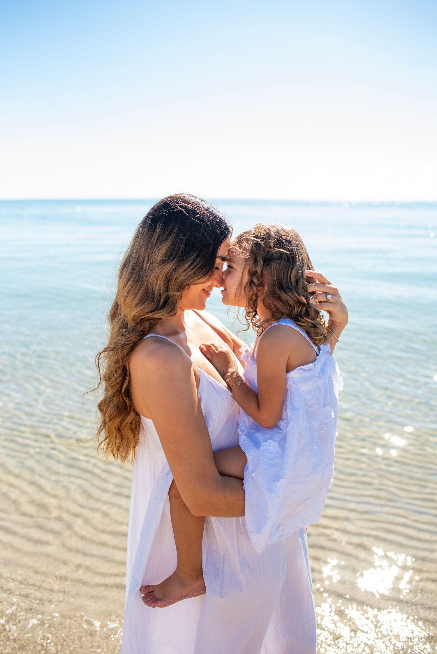 A tender moment shared between parent and child in white dresses on a sunny beach.