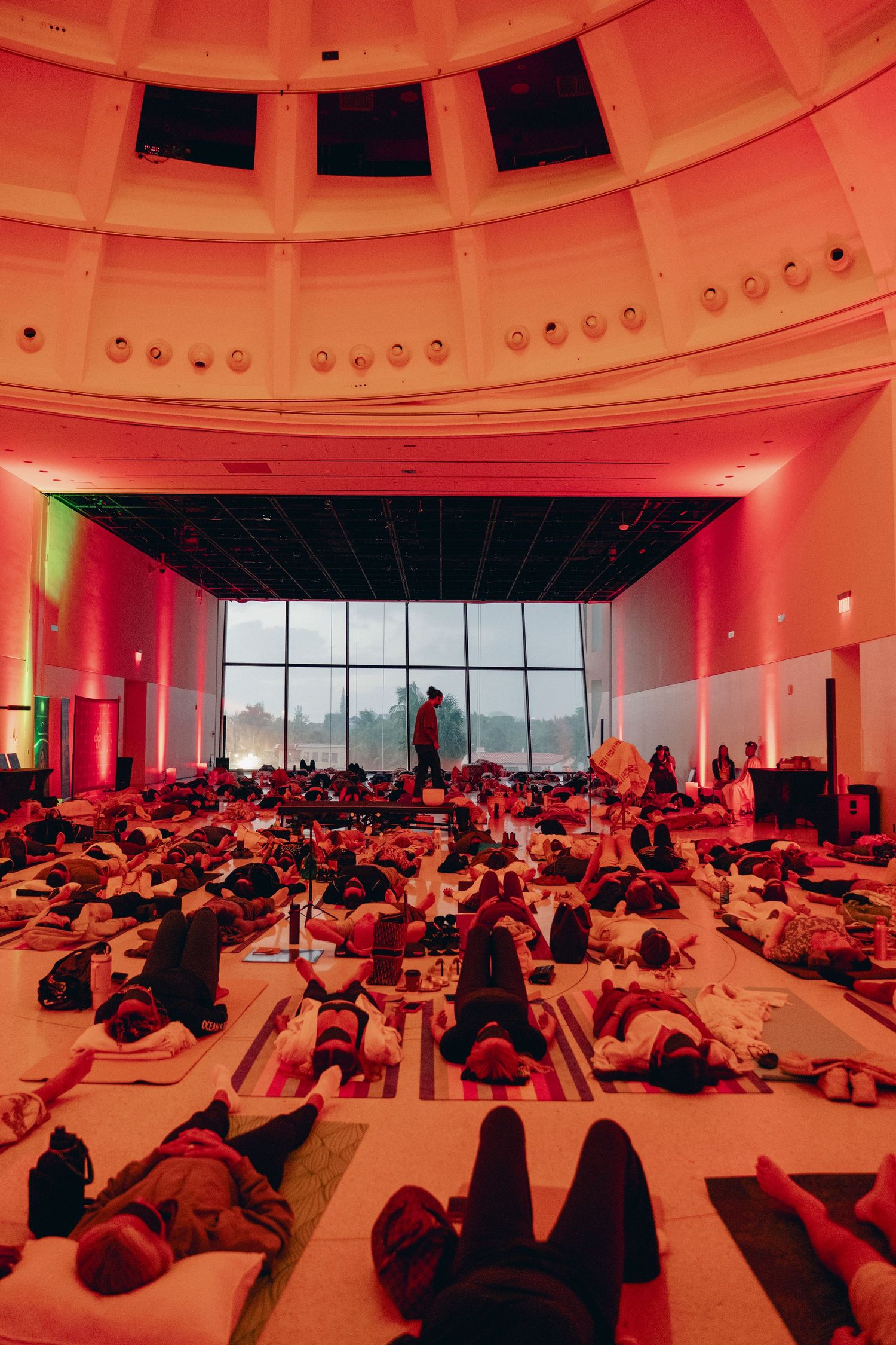 Large group yoga session in a domed hall bathed in red ambient lighting, participants lying on mats during relaxation pose.