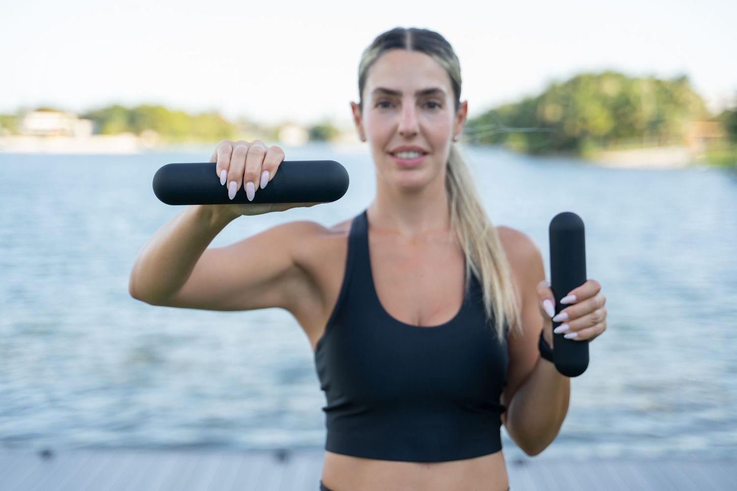 Woman in black workout attire holding gray exercise equipment by a waterfront.