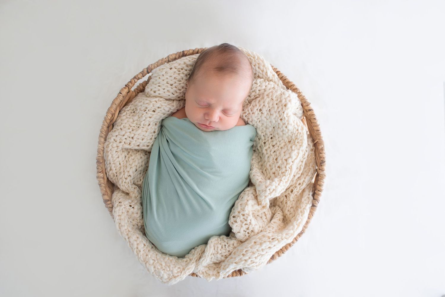 Newborn baby wrapped in mint green swaddle on cream blanket.