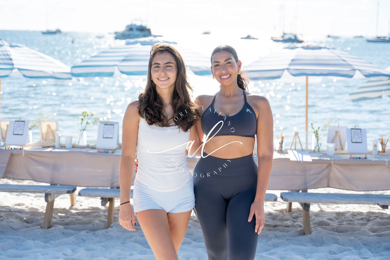 Two people smile together on a sunny beach with white umbrellas and lounge chairs in the background.