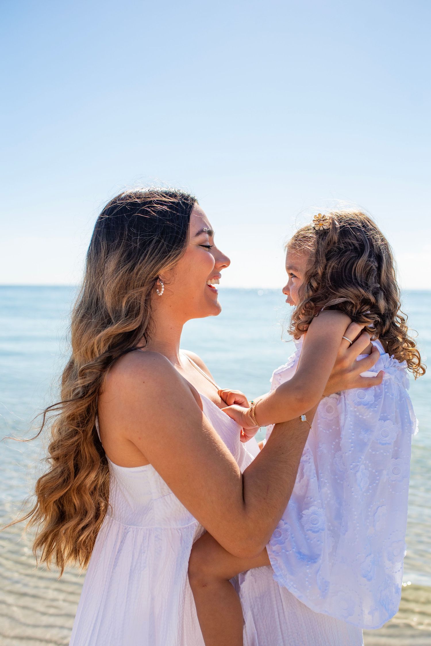 Mother and daughter sharing loving laughter while wearing matching white dresses on a sunny beach.