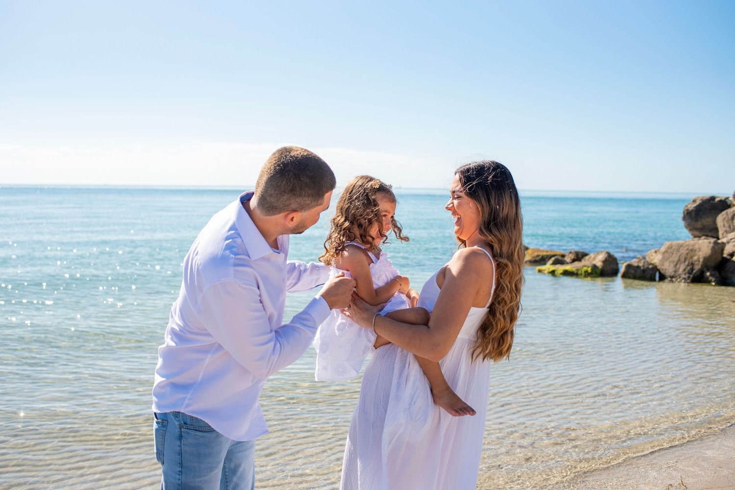 Couple embraces on a sunny beach with sparkling ocean waves and wooden pier in the background.