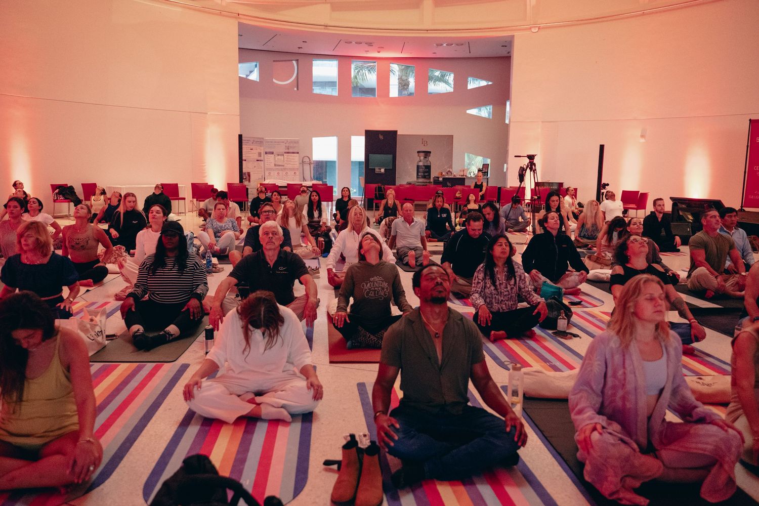 Large group of people sitting in meditation pose on colorful yoga mats in a spacious well-lit studio during a yoga event.
