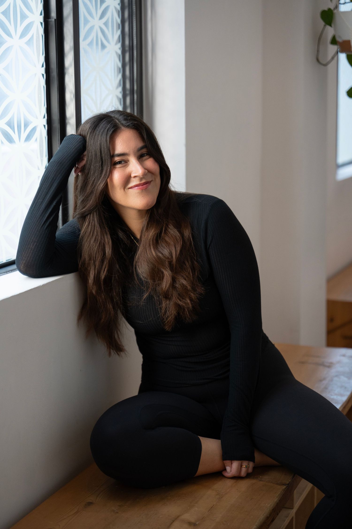 Person in black outfit sits and poses playfully on wooden floor near window with geometric pattern decor.