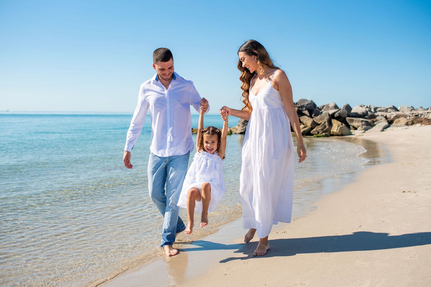 A family wearing white outfits walks together along a sunny beach with turquoise waters and rocky jetty in the background.
