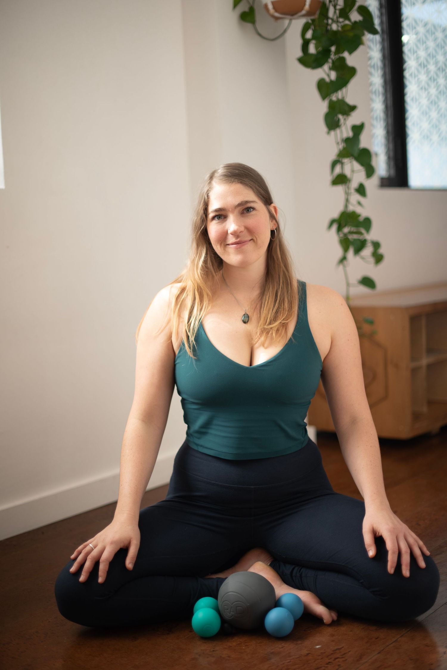 A person wearing a dark green top and black pants sits on a hardwood floor with a small green exercise ball.