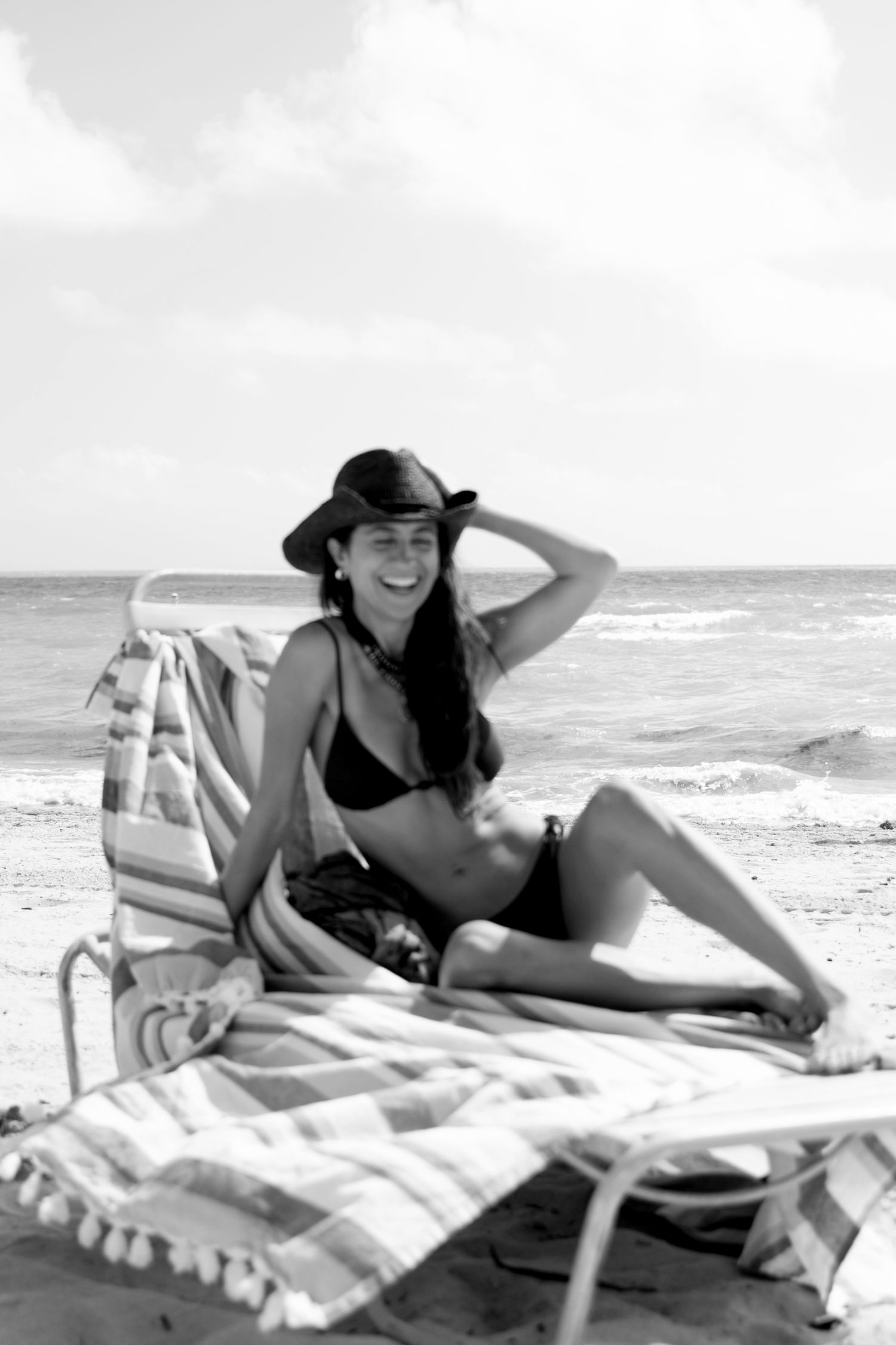 A woman in a dark swimsuit and sunhat relaxes on a striped beach lounger by the ocean.