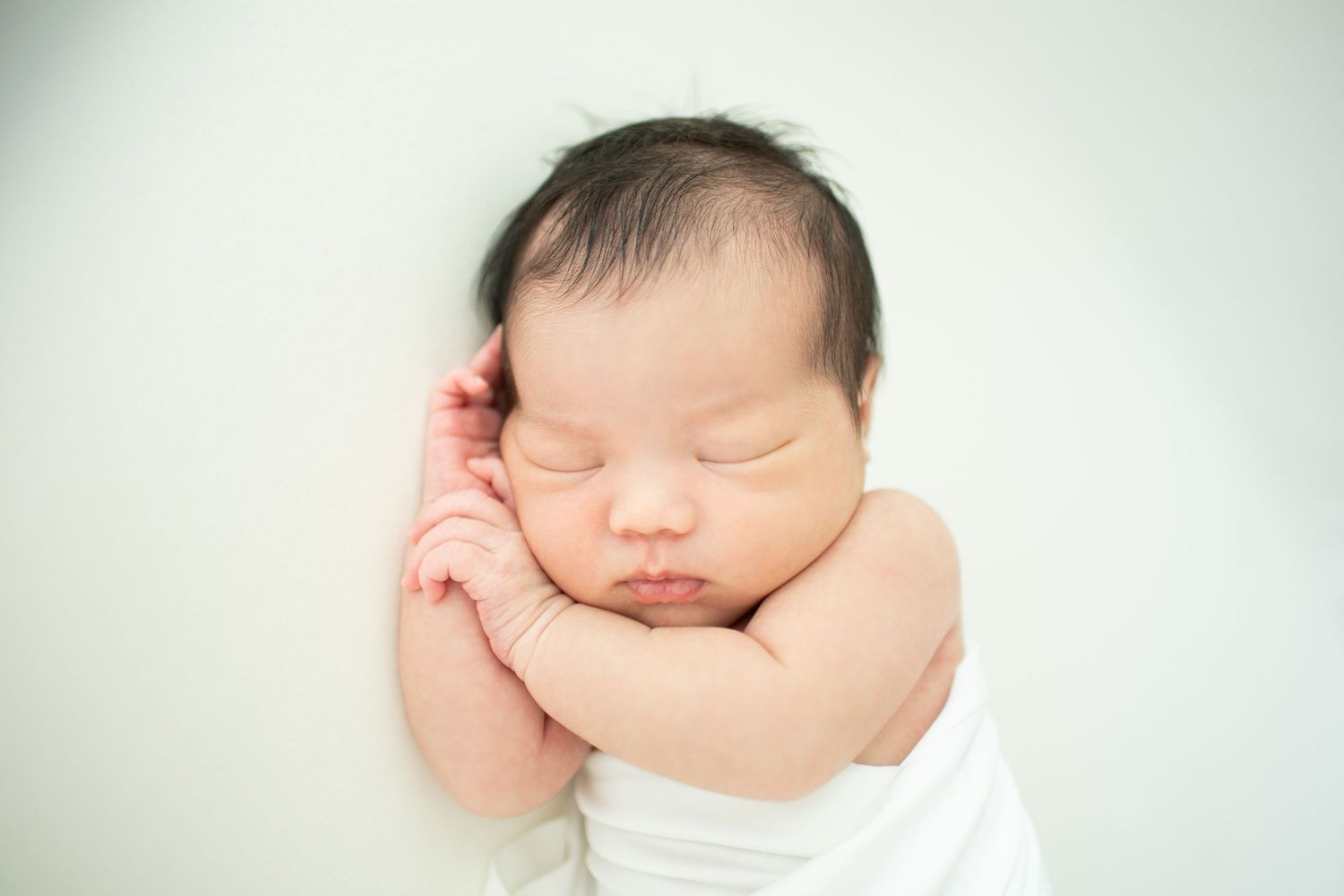 Peaceful sleeping newborn baby with chubby cheeks wearing white in soft focus portrait against light background.