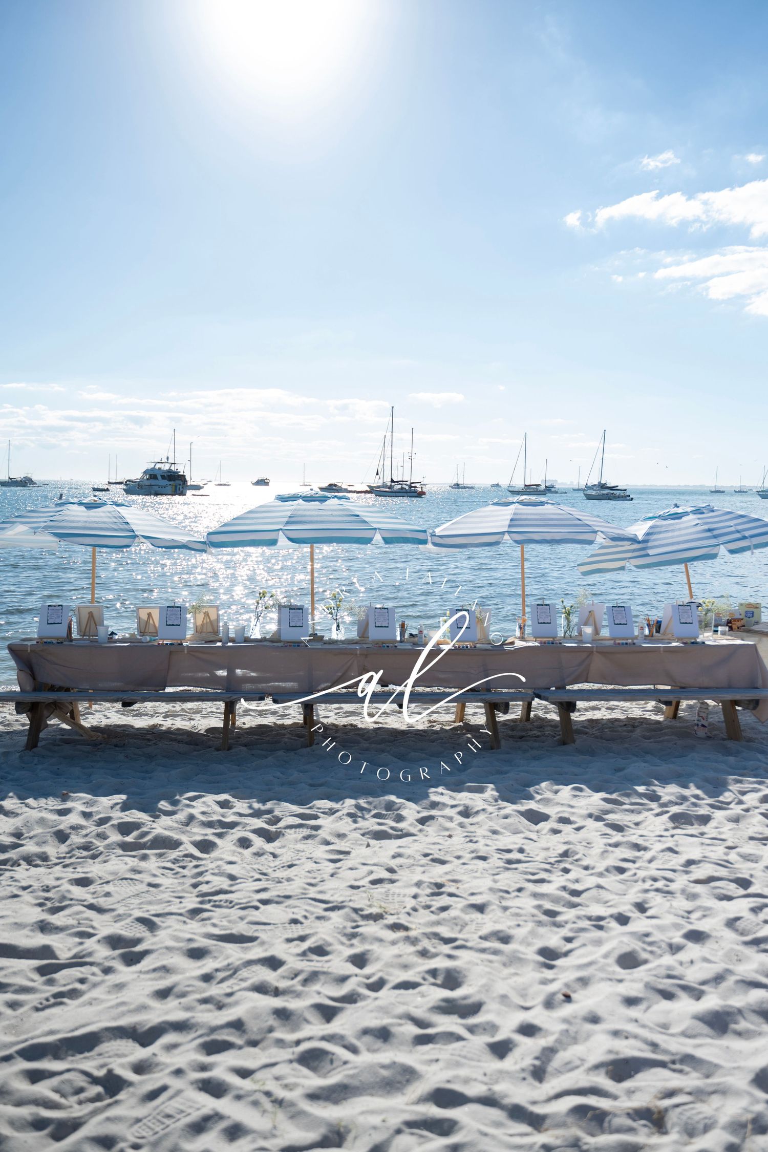 White beach umbrellas line a sandy shore with boats and yachts dotting the calm blue waters of a harbor on a sunny day.