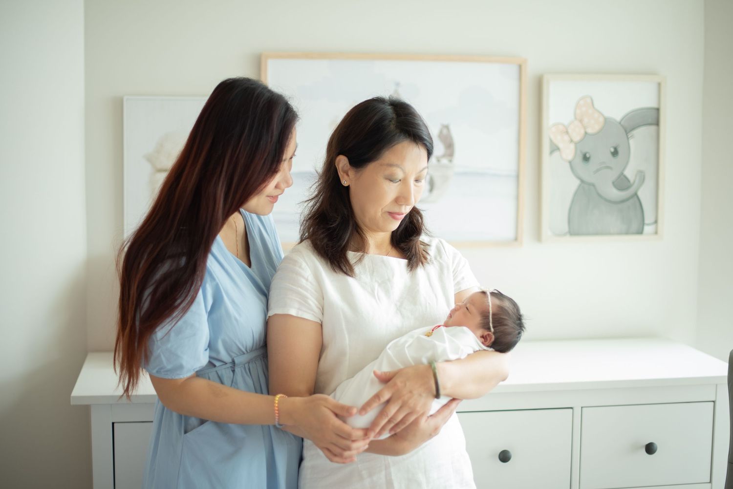 Two women stand in a nursery room looking lovingly at a newborn baby swaddled in white.