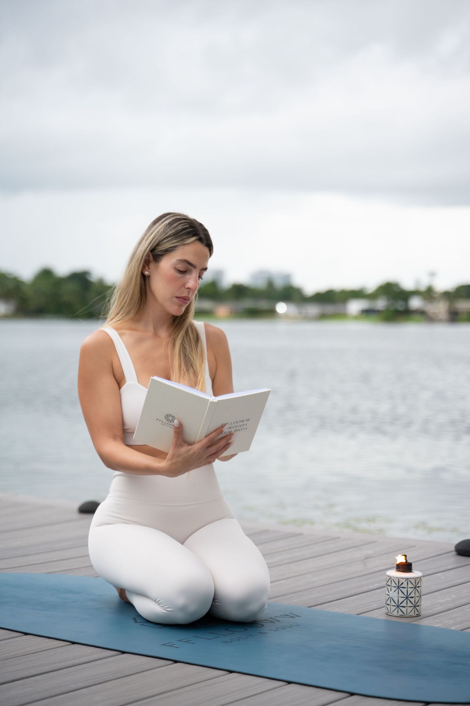Person in white athletic wear reading while seated on blue yoga mat by the lake.