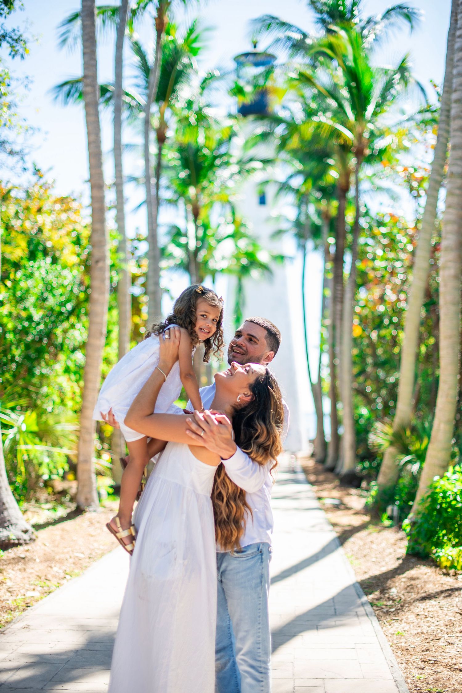 A romantic couple in white outfits shares intimate moments along a palm tree-lined pathway in a tropical setting.