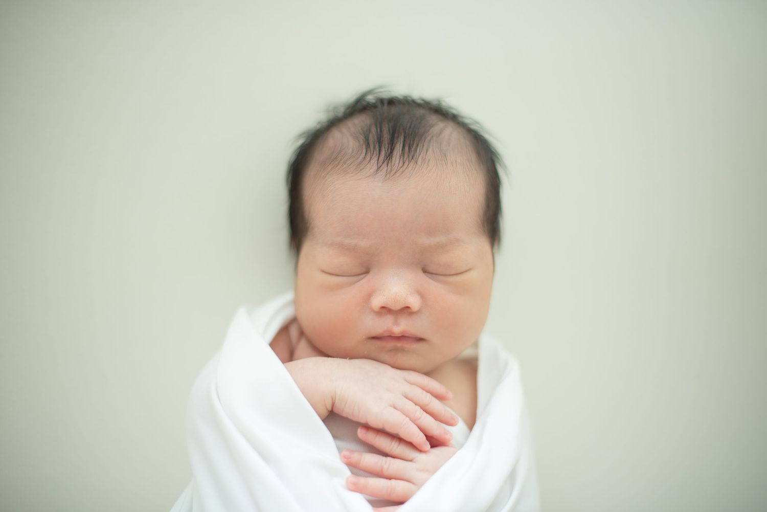 A sleeping newborn baby swaddled in white blankets rests peacefully against a light green background.